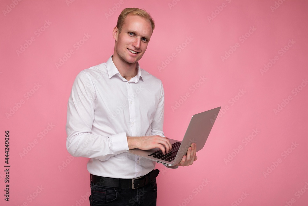 Side profile photo shot of handsome smiling happy confident blonde man holding and using computer laptop typing on keyboard wearing white shirt looking at camera isolated over pink background Side profile photo shot of handsome smiling happy confident blonde man holding and using computer laptop typing on keyboard wearing white shirt looking at camera isolated over pink background