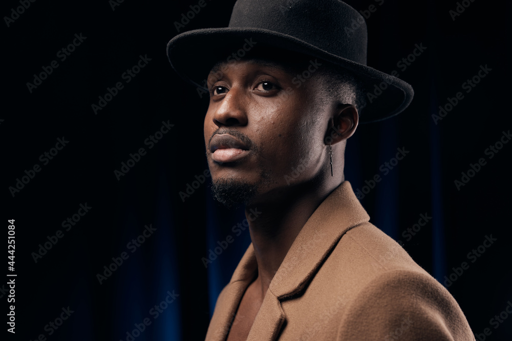 Obraz premium Portrait of a dark-skinned handsome man looking to the side. Dressed in a brown jacket and black hat. Portrait on black background