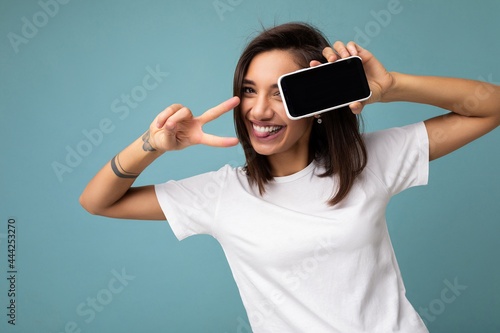 Attractive smiling young brunet woman good looking wearing white t-shirt standing isolated on blue background with copy space holding smartphone showing phone in hand with empty screen display for