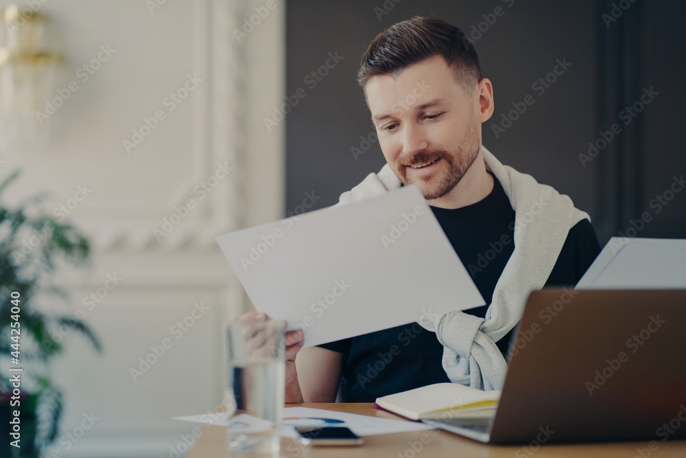 Indoor shot of male freelancer concentrated at papers prepares project or financial report poses at desktop with laptop computer works fron home blurred cozy background wears casual clothes.
