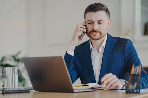 Serious businessman poses in office at desktop makes phone call concentrated at laptop display makes notes in diary plans working schedule dressed in formal clothes being busy. Digital management