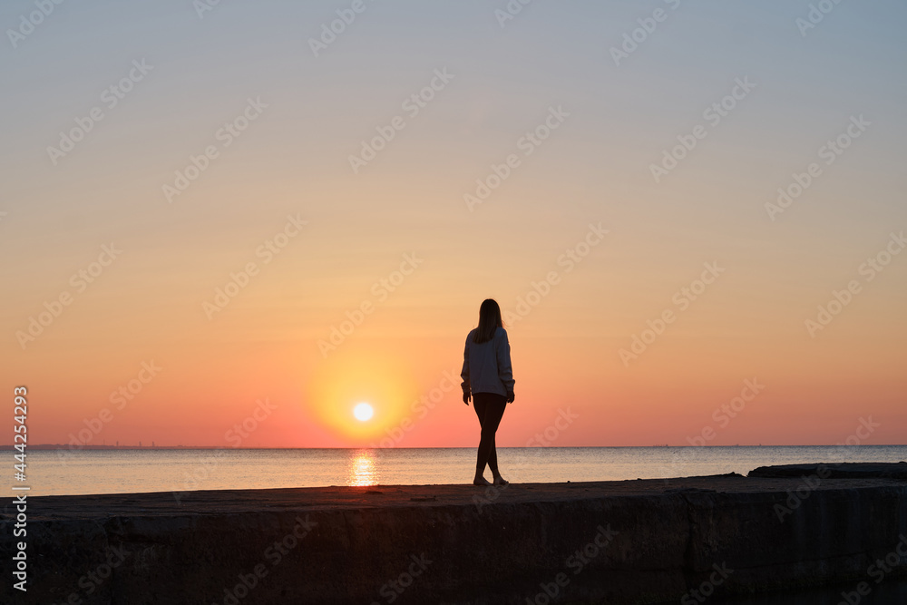 Beautiful girl stands by the sea and looks at the dawn. Girl in travel. Amazing scenic outdoors view. Beautiful dawn on the seashore. Adventure lifestyle