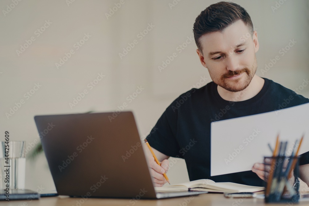 Skilled professional male expert busy doing paper report edits online files via laptop works at desktop focused at documents dressed in casual black t shirt. Bearded freelancer at home office Skilled professional male expert busy doing paper report edits online files via laptop works at desktop focused at documents dressed in casual black t shirt. Bearded freelancer at home office