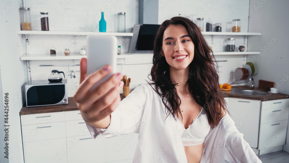 cheerful woman in bra and white shirt taking selfie on cellphone in kitchen cheerful woman in bra and white shirt taking selfie on cellphone in kitchen