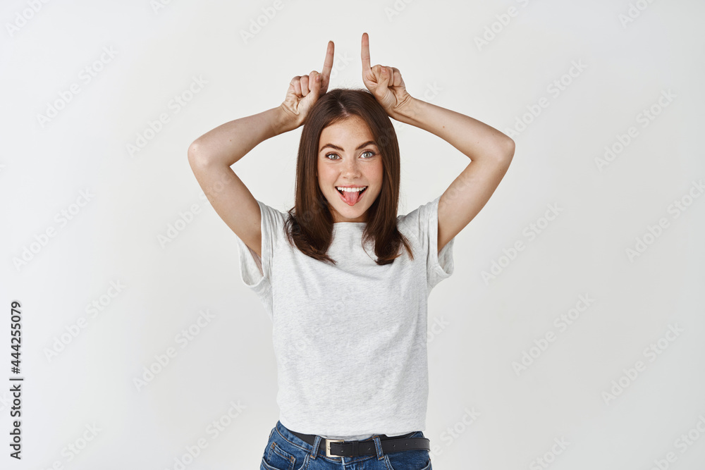 Girl showing inner devil. Portrait of charming cute and confident european female with tattoo on arm holding index fingers like demon horns on head and smiling with intrigued delighted face