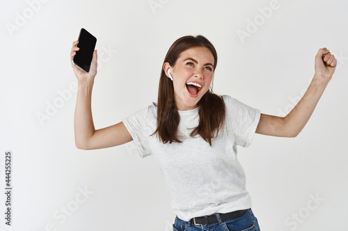 Happy woman listening music in wireless headphones and holding smartphone, dancing and singing over white background