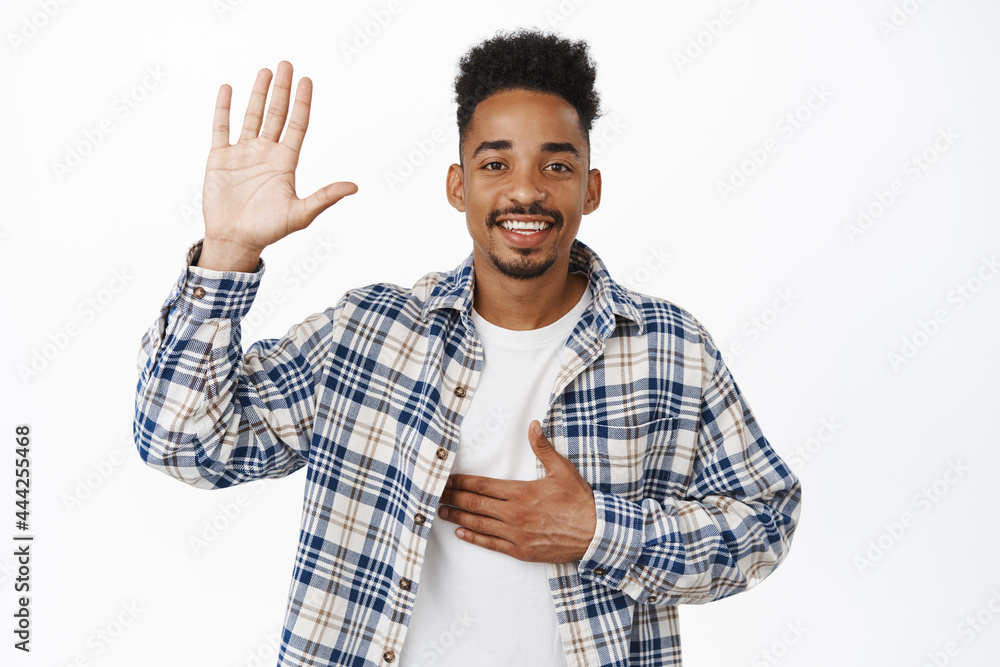 Greeting. Smiling friendly african american guy tell his name, raise hand and put arm on chest, introduce himself, say hello, making promise, standing over white background Greeting. Smiling friendly african american guy tell his name, raise hand and put arm on chest, introduce himself, say hello, making promise, standing over white background