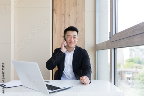 Happy young Asian business man in office looking at mobile phone, with emotion winner or win, financial stock sports betting. Male joyfully exclaims playing game. Excited overjoyed celebrating success