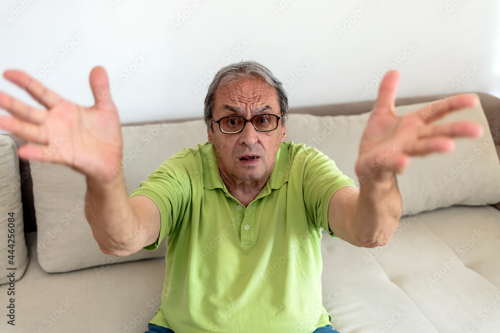 Naklejka premium Photo of a old man arguing, shouting, angry and emotionally gesturing at home. Irritated senior man in casual clothes at home wear glasses, yelling while sitting at home isolated on bright background.