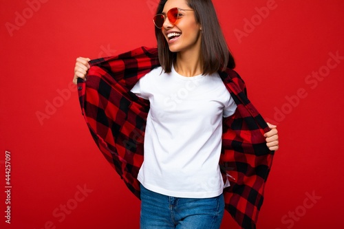Photo of young beautiful happy smiling brunette woman wearing trendy white t-shirt with empty space for mock up and red check shirt and red sunglasses. Sexy carefree female person posing isolated near