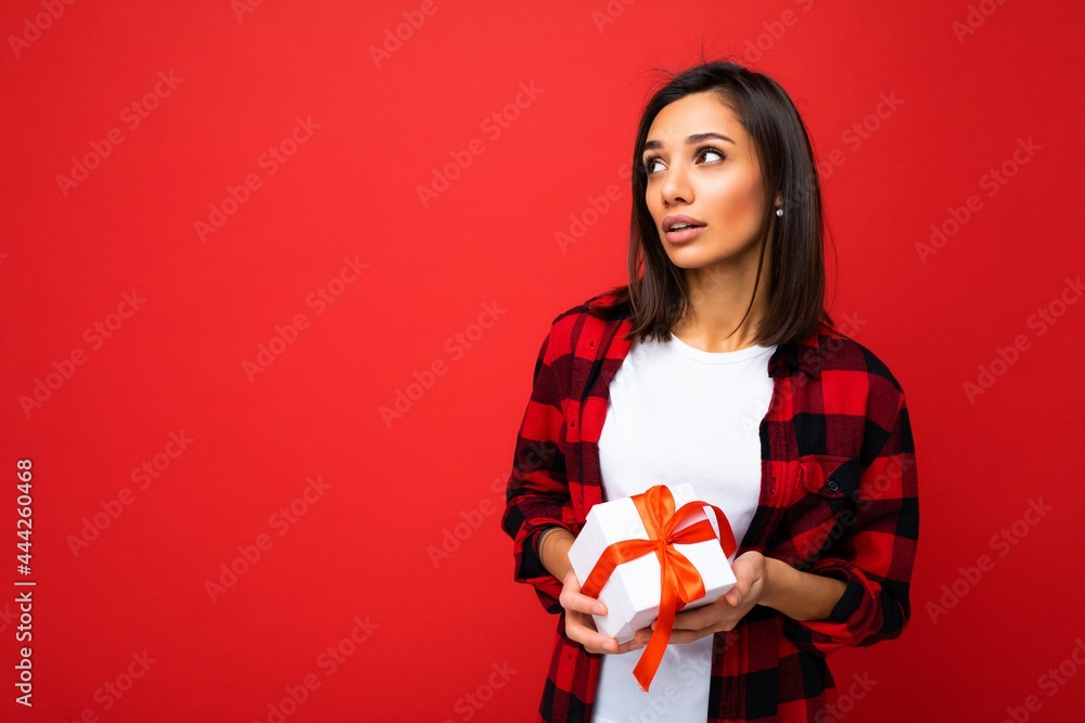 Photo shot of pretty positive thoughtful young brunette woman isolated over red background wall wearing white casual t-shirt and red and black shirt holding white gift box with red ribbon and looking Photo shot of pretty positive thoughtful young brunette woman isolated over red background wall wearing white casual t-shirt and red and black shirt holding white gift box with red ribbon and looking
