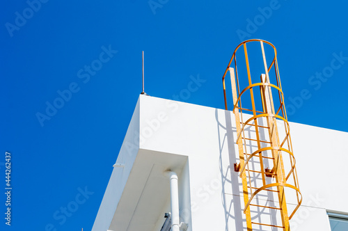 yellow metal ladder to roof (fire escape) on a white wall of building,safety ladder for roof maintenance or fire emergency escape ladder on a building vertical in angle,blue sky in background.