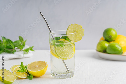 Sweet organic lemonade summer cold drink made of sour ripe lemon and lime slices, carbonated water and mint leaves served in drinking glass with straw on white wooden table with ingredients at kitchen
