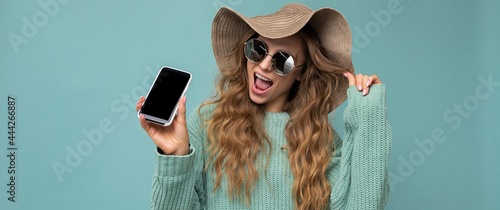 Panoramic photo of beautiful happy young blonde woman wearing sunglasses and summer hat isolated on blue background with copy space holding smartphone showing phone in hand with empty screen display
