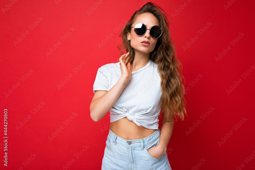 Photo shot of beautiful young dark blonde woman wearing casual clothes and stylish sunglasses isolated over colorful background looking at camera Photo shot of beautiful young dark blonde woman wearing casual clothes and stylish sunglasses isolated over colorful background looking at camera
