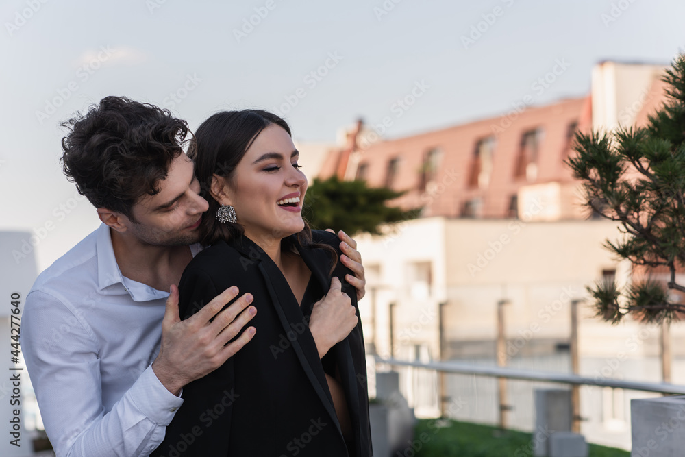 man hugging cheerful woman in black blazer on terrace man hugging cheerful woman in black blazer on terrace
