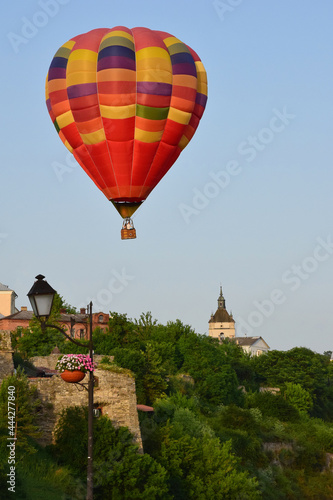 A large multicolored hot air balloon flies low over ancient buildings and a tree-lined valley. Photographed at close range