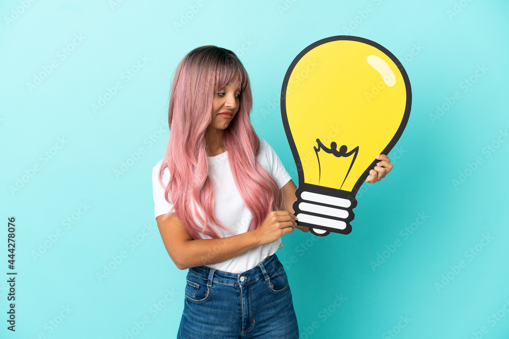 Young mixed race woman with pink hair isolated on blue background holding a bulb icon and having doubts Young mixed race woman with pink hair isolated on blue background holding a bulb icon and having doubts