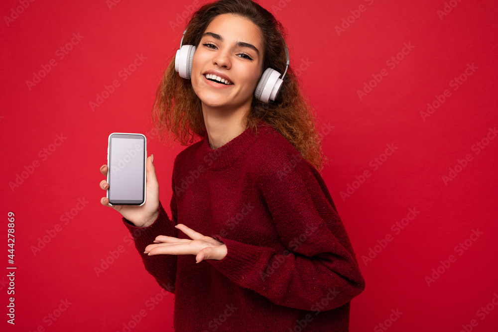 Attractive positive smiling young woman wearing stylish casual outfit isolated on colourful background wall holding and showing mobile phone with empty screen for cutout wearing white bluetooth Attractive positive smiling young woman wearing stylish casual outfit isolated on colourful background wall holding and showing mobile phone with empty screen for cutout wearing white bluetooth