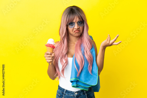 Young mixed race woman with pink hair holding ice cream isolated on yellow background having doubts while raising hands