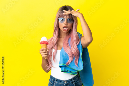 Young mixed race woman with pink hair holding ice cream isolated on yellow background doing surprise gesture while looking to the side