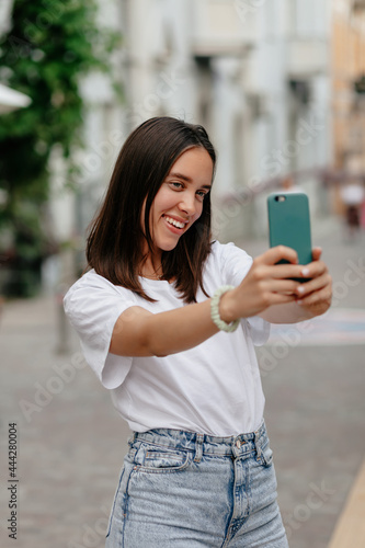 Charming adorable lady with short hairstyle wearing white t-shirt making selfie with smartphone while walking on the street . High quality photo
