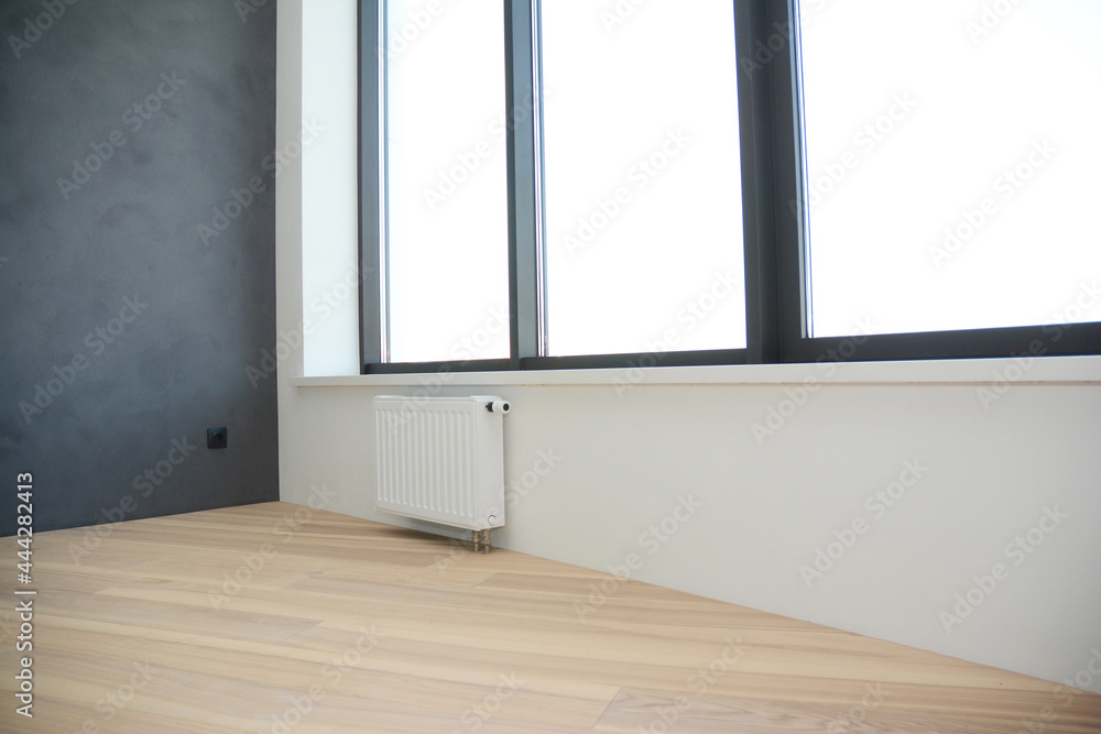 An empty modern apartment with oak hardwood flooring, gray plastered wall, large windows and a radiator. An empty modern apartment with oak hardwood flooring, gray plastered wall, large windows and a radiator.