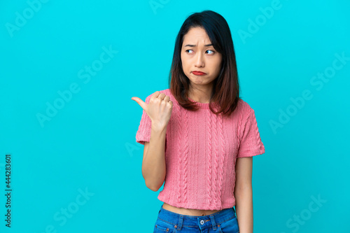 Young Vietnamese woman isolated on blue background unhappy and pointing to the side