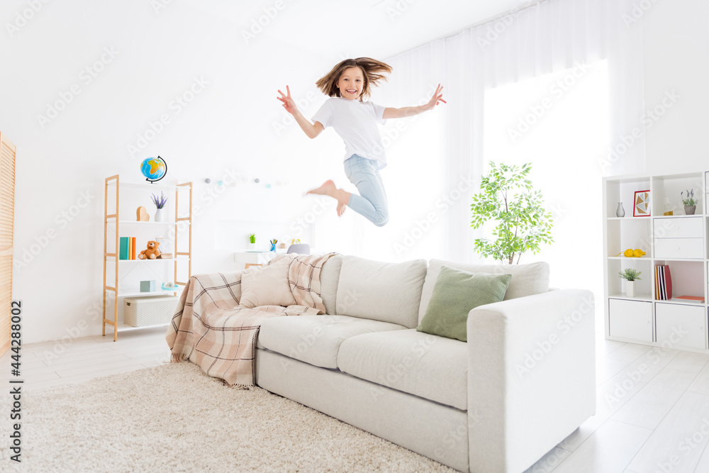 Photo of sweet charming school girl wear white t-shirt smiling jumping sofa showing v-signs indoors house home room Photo of sweet charming school girl wear white t-shirt smiling jumping sofa showing v-signs indoors house home room
