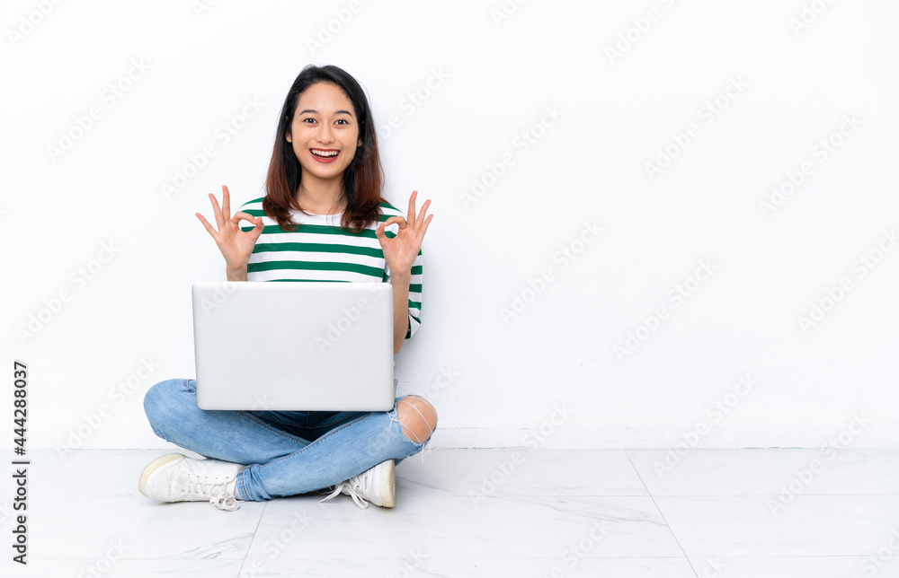 Naklejka premium Young Vietnamese woman with a laptop sitting on the floor isolated on white wall showing an ok sign with fingers