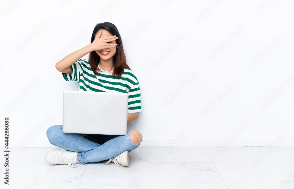 Young Vietnamese woman with a laptop sitting on the floor isolated on white wall covering eyes by hands and smiling Young Vietnamese woman with a laptop sitting on the floor isolated on white wall covering eyes by hands and smiling