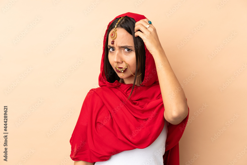 Young Indian woman isolated on beige background having doubts while scratching head Young Indian woman isolated on beige background having doubts while scratching head