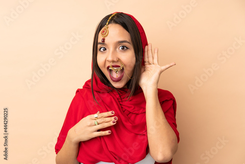 Young Indian woman isolated on beige background listening to something by putting hand on the ear