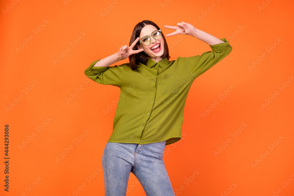 Photo of crazy brunette hairdo young lady show v-sign wear specatcles green shirt isolated on orange color background Photo of crazy brunette hairdo young lady show v-sign wear specatcles green shirt isolated on orange color background