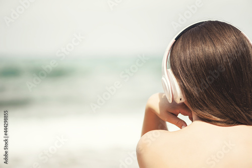 Back view of alone woman sitting on a beach in headphones listen music dreaming and looking on the sea. Female relaxation at summer vacation.