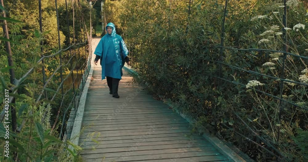 Woman in blue raincoat is engaged in hiking. On summer evening at sunset, she walks along wooden suspension bridge over river. The lady smiles. Concept of savage travel, lifestyle, outdoor recreation Woman in blue raincoat is engaged in hiking. On summer evening at sunset, she walks along wooden suspension bridge over river. The lady smiles. Concept of savage travel, lifestyle, outdoor recreation
