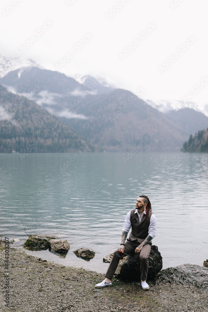 A brutal thoughtful young man groom with a dreadlocked hairstyle in a fashionable elegant wedding suit stands on the shore of a lake against the background of misty mountains in nature A brutal thoughtful young man groom with a dreadlocked hairstyle in a fashionable elegant wedding suit stands on the shore of a lake against the background of misty mountains in nature