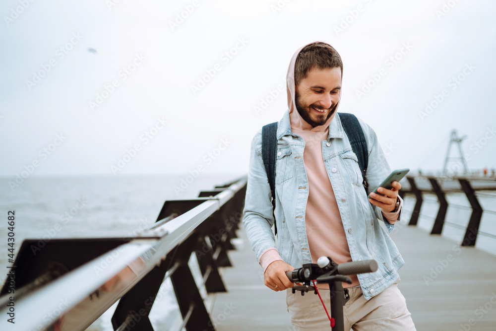 Young man with phone riding electric scooter on a pier near the sea. Hipster man taking pictures on phone during travel. ​Selfie time, blogging. Ecological transportation concept.