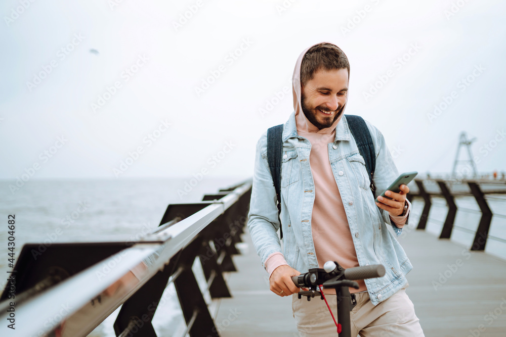 Young man with phone riding electric scooter on a pier near the sea. Hipster man taking pictures on phone during travel. Selfie time, blogging. Ecological transportation concept. Young man with phone riding electric scooter on a pier near the sea. Hipster man taking pictures on phone during travel. Selfie time, blogging. Ecological transportation concept.