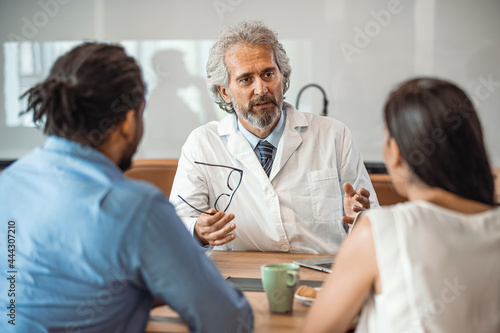 Doctor speaking to young couple. Doctor Using Tablet Computer Discussing Treatment With Patients. Mature Doctor Involved In Serious Discussion With His Patients