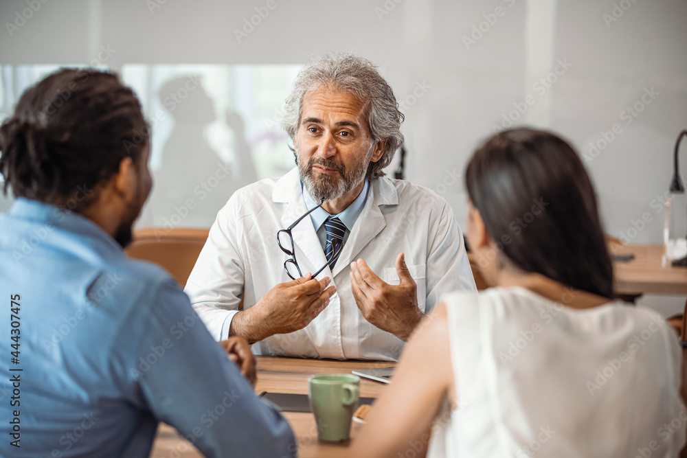 Male doctor offering medical advices to a young couple in office. Husband and wife in a clinic, Beautiful young couple at pregnancy consultation period. Couple meeting medical specialist at hospital Male doctor offering medical advices to a young couple in office. Husband and wife in a clinic, Beautiful young couple at pregnancy consultation period. Couple meeting medical specialist at hospital