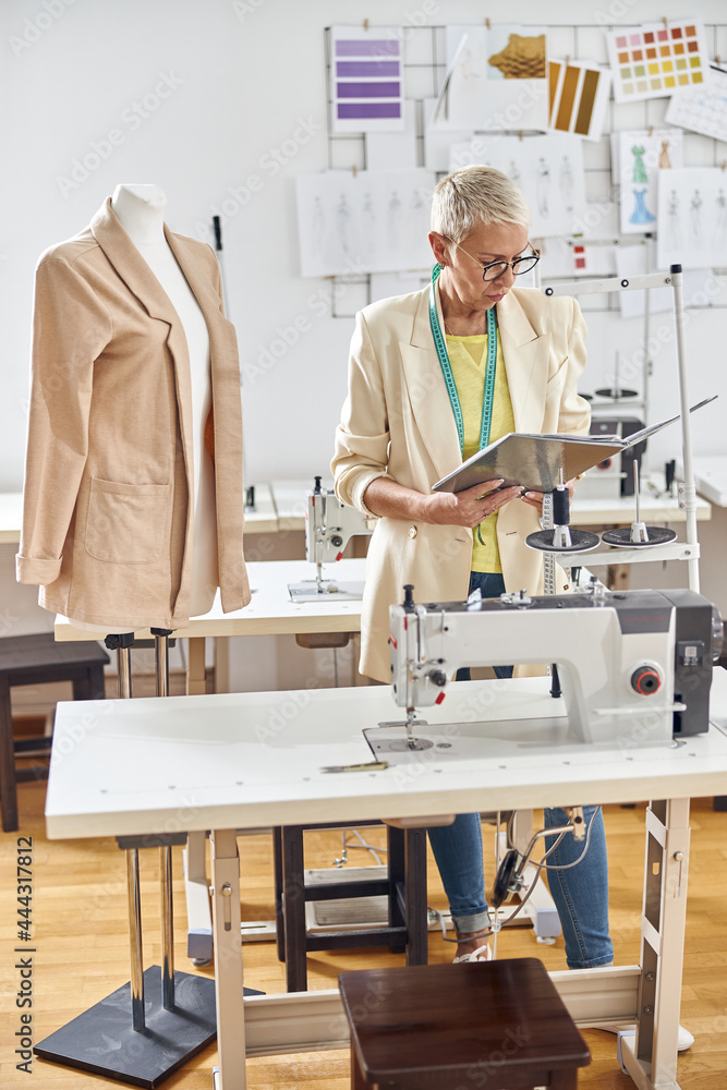 Middle aged fashion designer looks at open folder standing by table with sewing machine Middle aged fashion designer looks at open folder standing by table with sewing machine