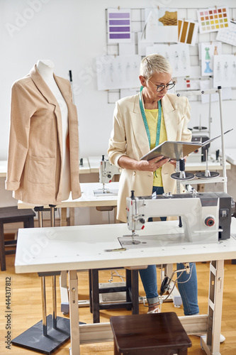 Middle aged fashion designer looks at open folder standing by table with sewing machine
