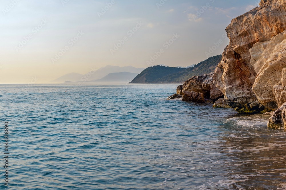 Beautiful landscape - beach in sunset sunlight - waving sea water, rocks and cliffs, blue sky and mountains on the horizon.