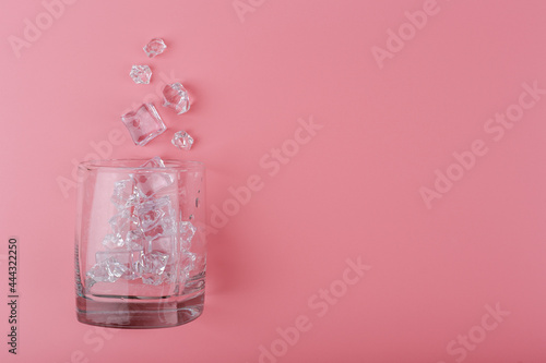 Creative flat lay with empty transparent glass with spilled ice cubes on pink background with copy space. Concept of summer hot, refreshing iced summer drinks and cold cocktails