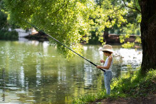 A boy with curly hair in a straw hat stands with a fishing rod by the pond. Child fishing on a summer day.