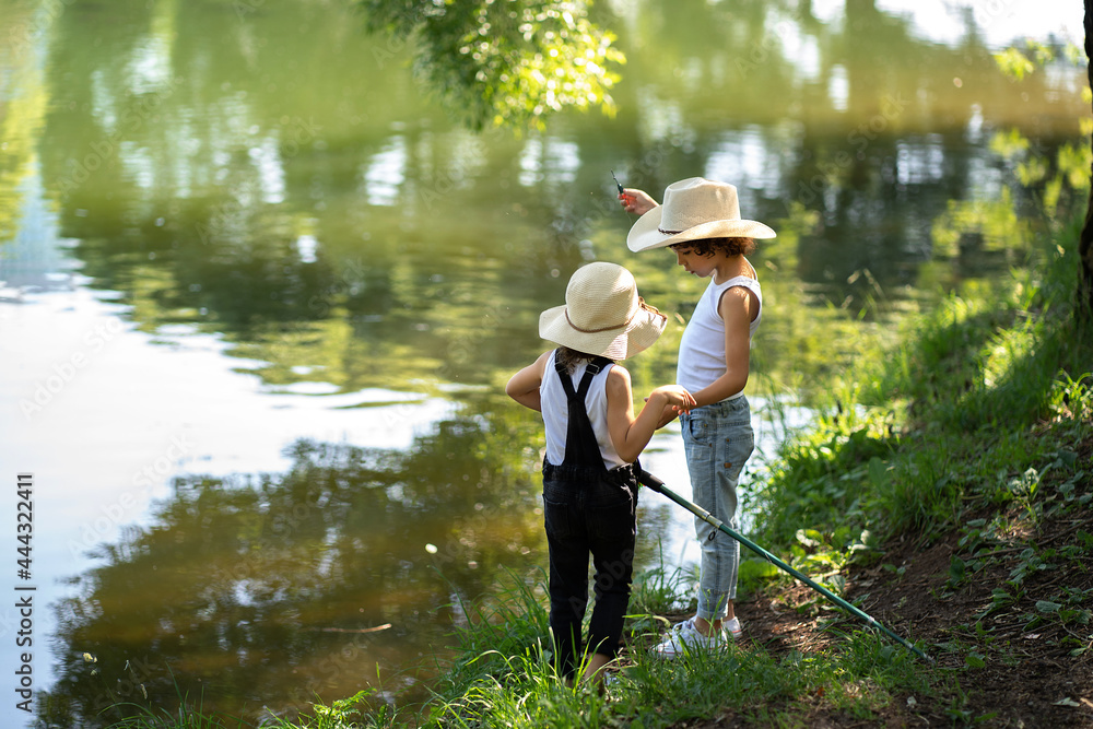 A boy and a girl with curly hair in straw hats stand with a fishing rod by the pond. Children fishing on a summer day. A boy and a girl with curly hair in straw hats stand with a fishing rod by the pond. Children fishing on a summer day.