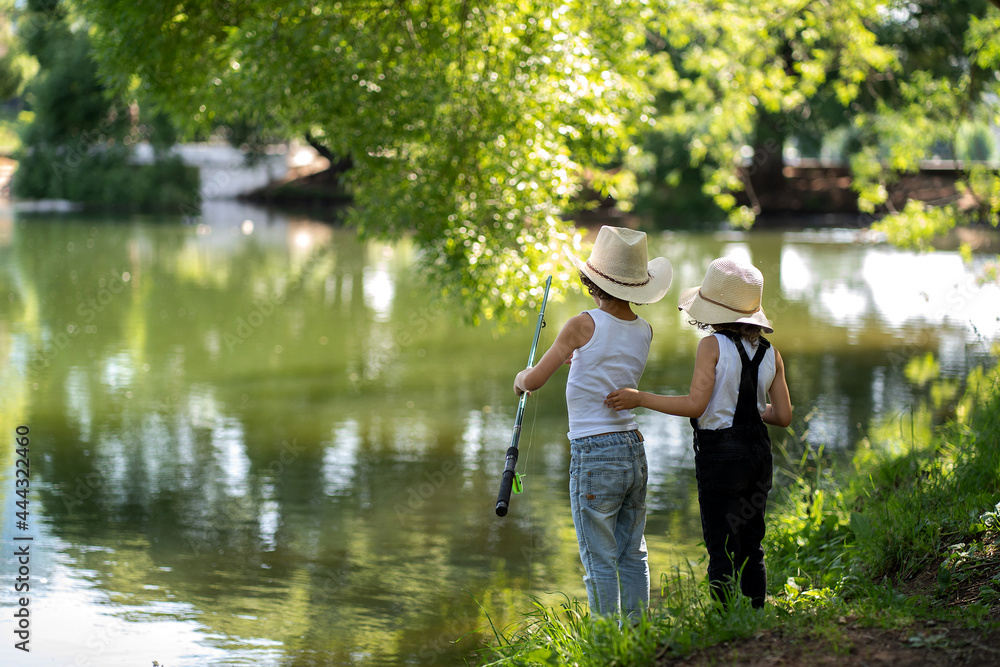 A boy and a girl with curly hair in straw hats stand with a fishing rod by the pond. Children fishing on a summer day. A boy and a girl with curly hair in straw hats stand with a fishing rod by the pond. Children fishing on a summer day.