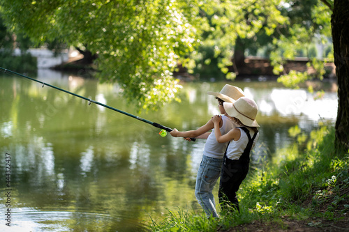 A boy and a girl with curly hair in straw hats stand with a fishing rod by the pond. Children fishing on a summer day.