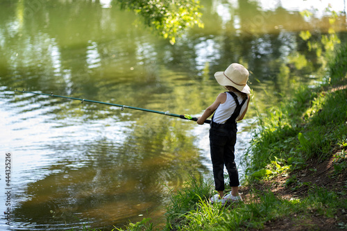A little girl with curly hair in a straw hat and black overalls stands with a fishing rod by the pond. Child fishing on a summer day